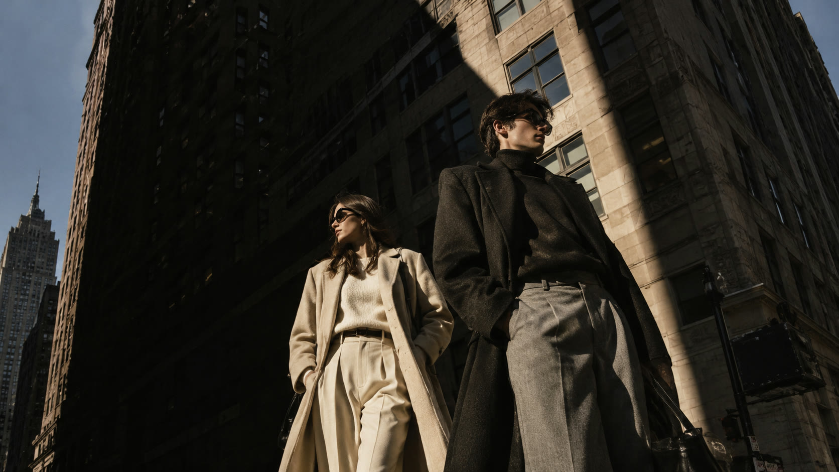 A woman in a cream coat and a man in a long black overcoat on a New York street, in a shaft of sunlight between tall buildings